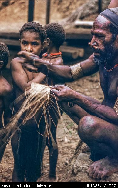 Papua New Guinean man weaving cane armband, children in background