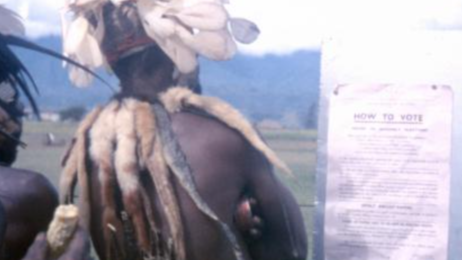 Man in traditional dress with feather headdress reading election sign
